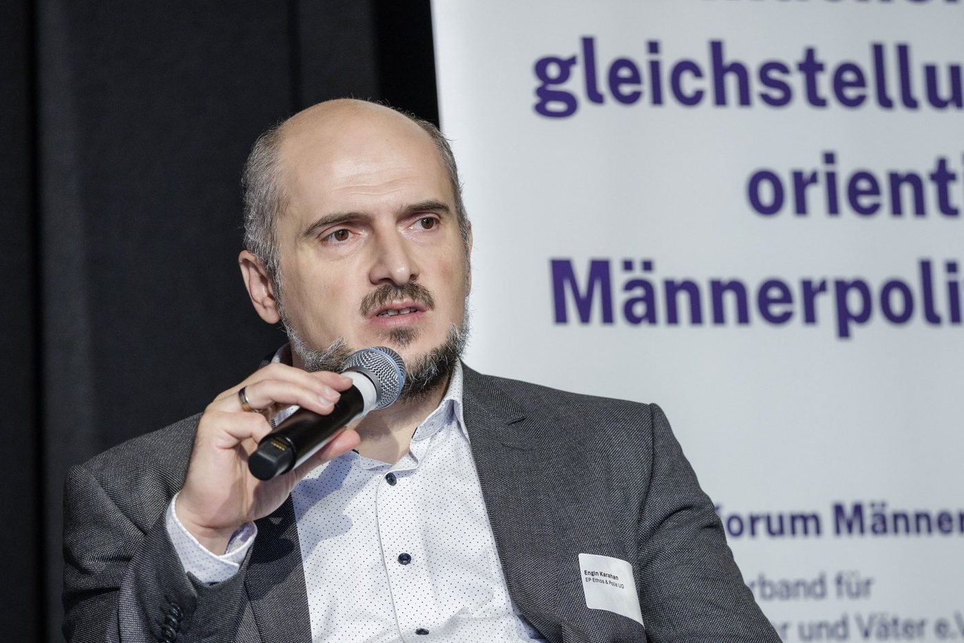 Engin Karahan spricht auf einem Podium in ein Mikrofon, hinter ihm ein Banner des Bundesforums Männer
