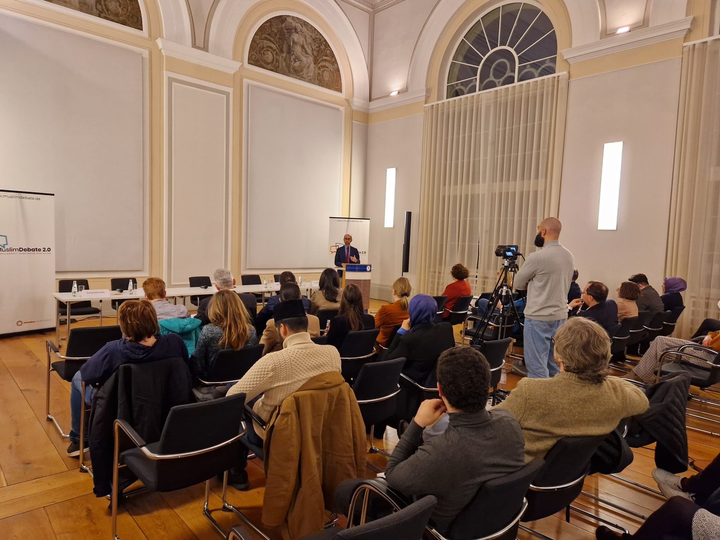 Publikum in einem hellen Festsaal, vorne ein Redner am Pult, rechts ein Kameramann an einer Videokamera