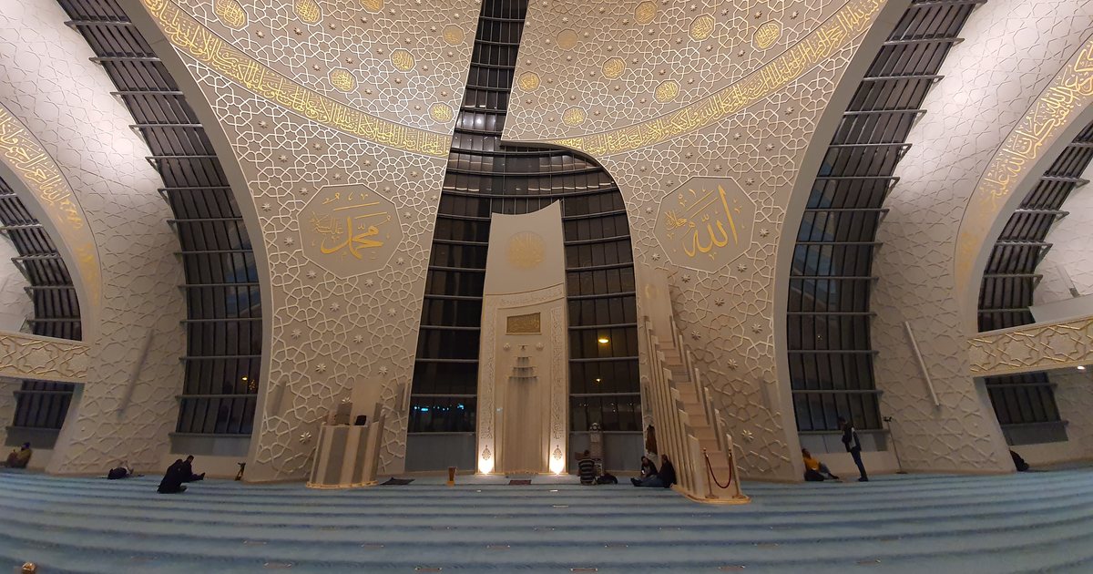 Gebetsraum der DITIB-Zentralmoschee in Köln mit Mihrab und Minbar unter der Kuppel, an den Wänden Kalligrafie, auf dem Teppich wenige Betende.
