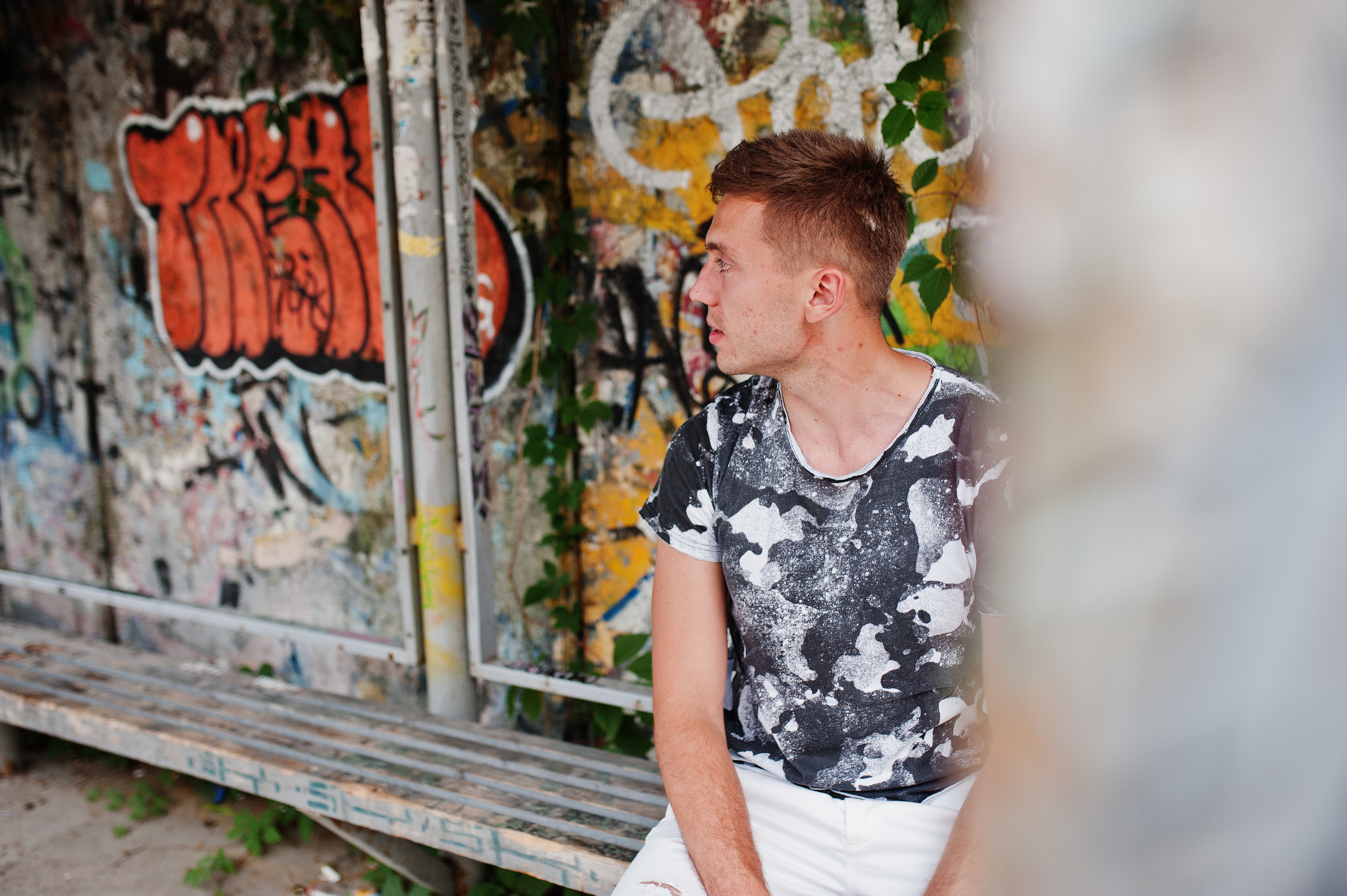 Junger Mann sitzt vor einer mit Graffiti bedeckten Wand in einem urbanen Umfeld und schaut nachdenklich zur Seite.