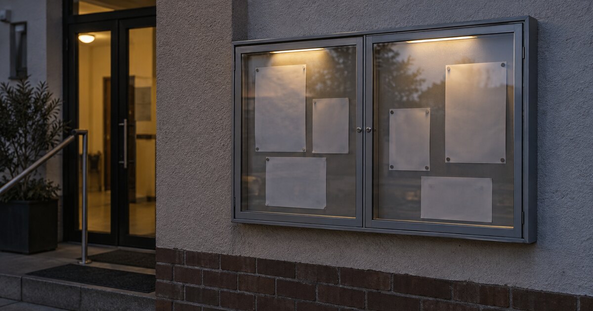 Beleuchteter Schaukasten mit zwei getrennten Aushangbereichen an der Außenwand eines Gemeindegebäudes im Abendlicht, die Aushänge sind unbeschriftet.