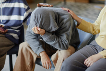 Drei Personen in einem Innenraum. Die Person in der Mitte sitzt mit gesenktem Kopf und verdecktem Gesicht, zwei andere legen ihr tröstend die Hand auf Schulter und Rücken.