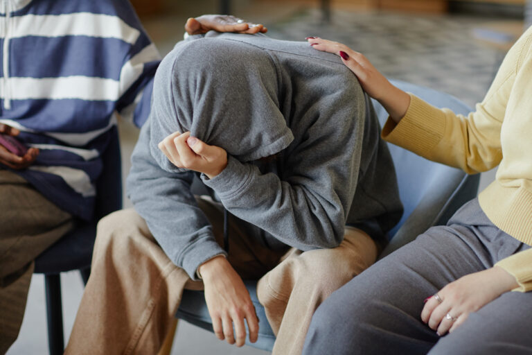 Drei Personen in einem Innenraum. Die Person in der Mitte sitzt mit gesenktem Kopf und verdecktem Gesicht, zwei andere legen ihr tröstend die Hand auf Schulter und Rücken.
