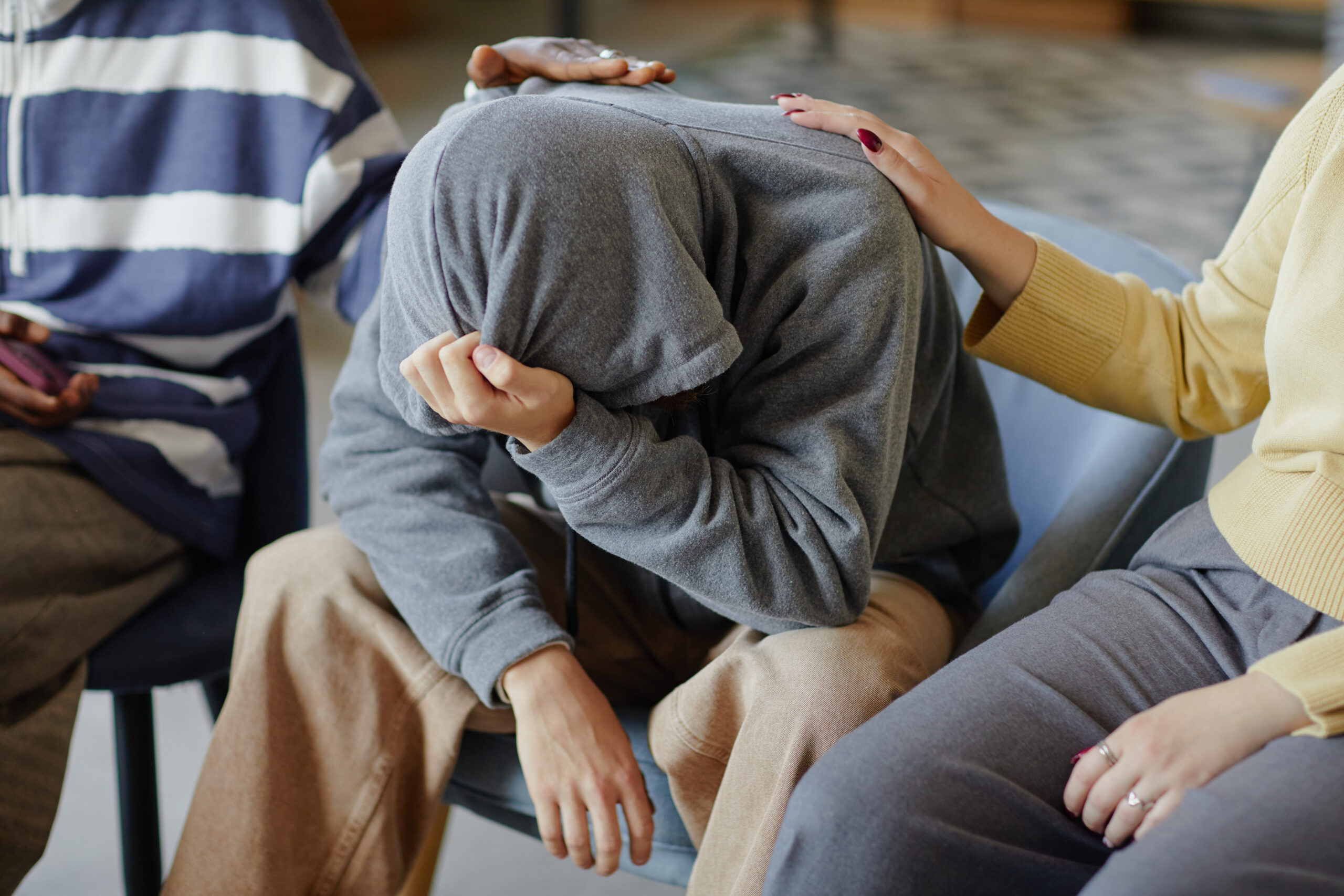 Drei Personen in einem Innenraum. Die Person in der Mitte sitzt mit gesenktem Kopf und verdecktem Gesicht, zwei andere legen ihr tröstend die Hand auf Schulter und Rücken.